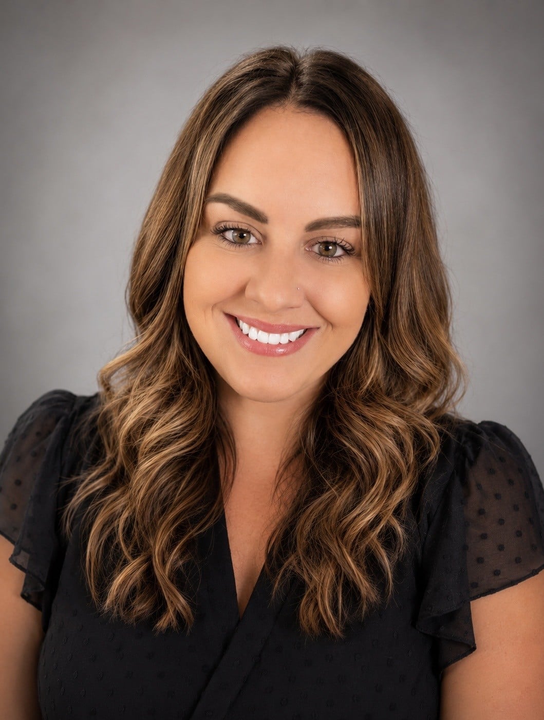 Professional headshot of a smiling woman with wavy brown hair in a black dotted blouse against a gray background.
