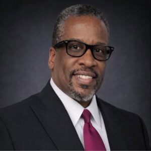5bcd2488-4fbd-437e-b4ca-855705415a7d | Portage Path Behavioral Health Professional headshot of a smiling man in a black suit, white shirt, and magenta tie, wearing glasses against a dark backdrop.