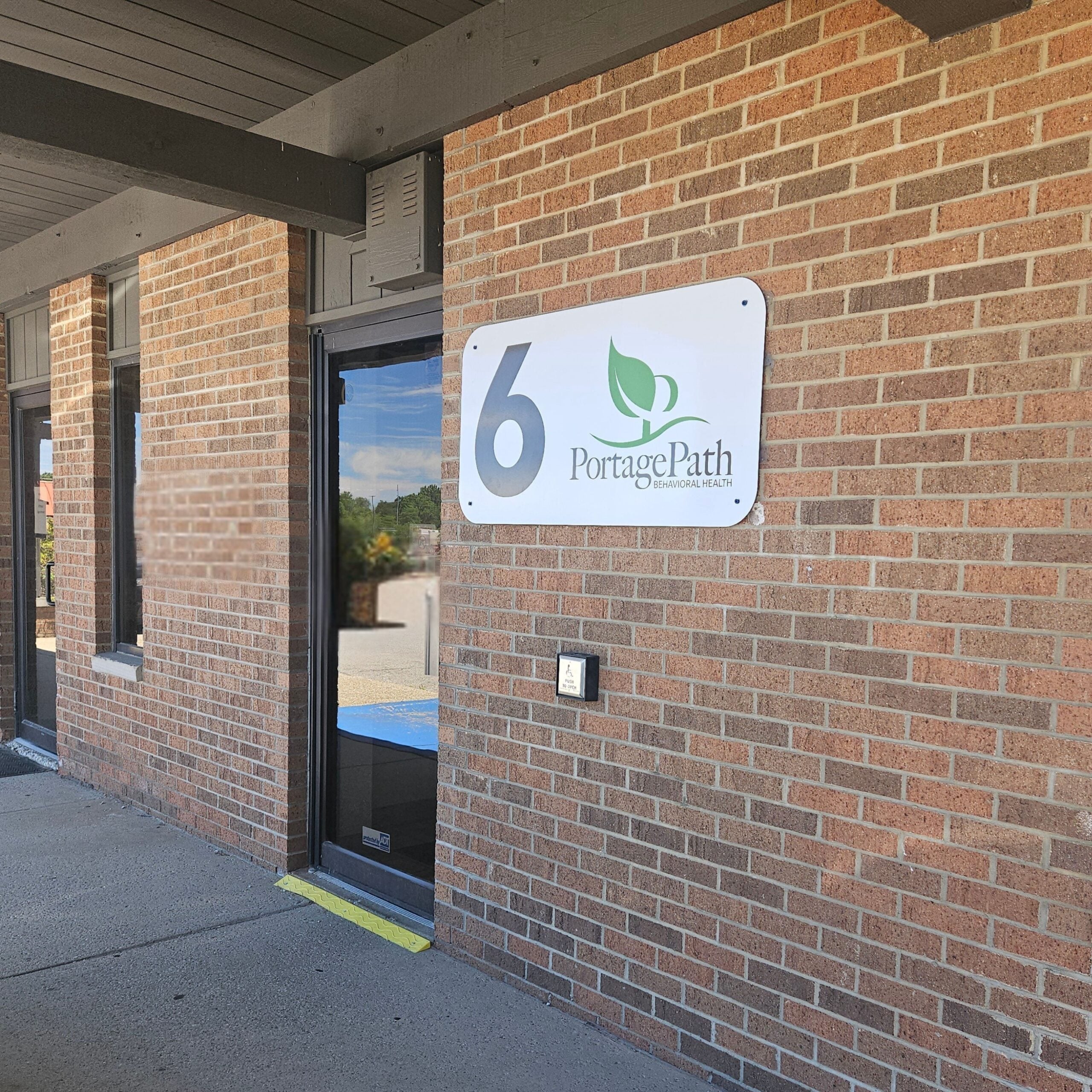 Barberton Front Door | Portage Path Behavioral Health Brick storefront entrance with a glass door and a portage path behavioral health sign on the brick wall nearby. It includes a yellow tactile ground strip at the doorway edge.