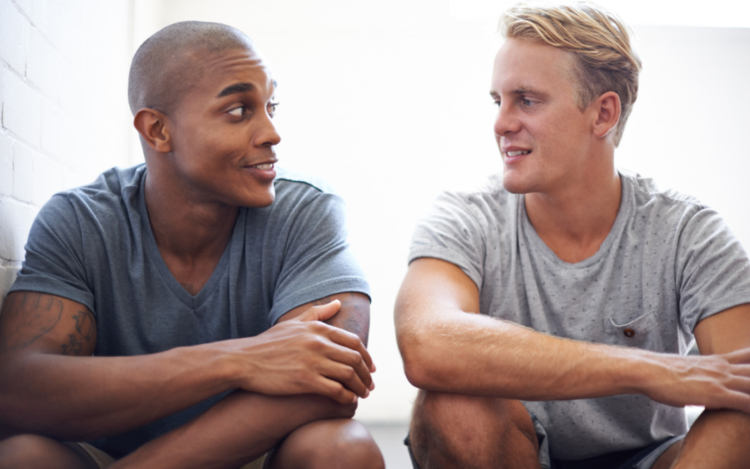 Two friends sitting side by side against a white brick wall, turned toward each other and smiling during a supportive conversation.