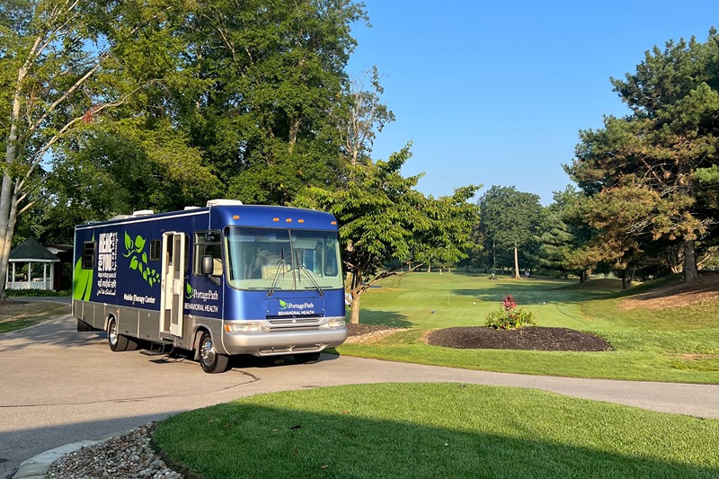 Img_1884 1 | Portage Path Behavioral Health Blue mobile clinic bus parked along a paved road beside a grassy, landscaped area with trees in the background.