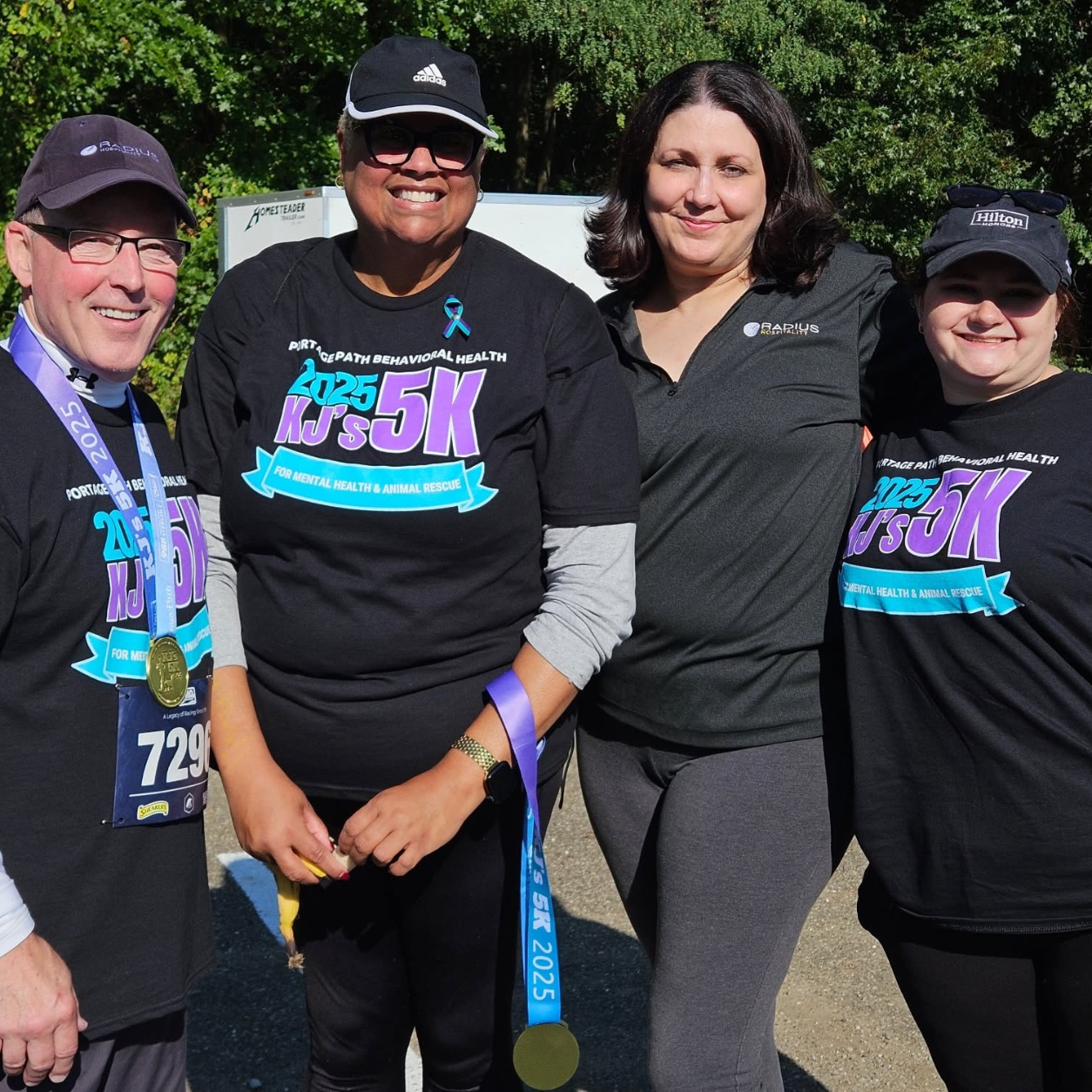 Four people pose together at an outdoor charity 5k, wearing matching black shirts with 'kj's 5k' logos and race medals visible. (informative group portrait after the race. )