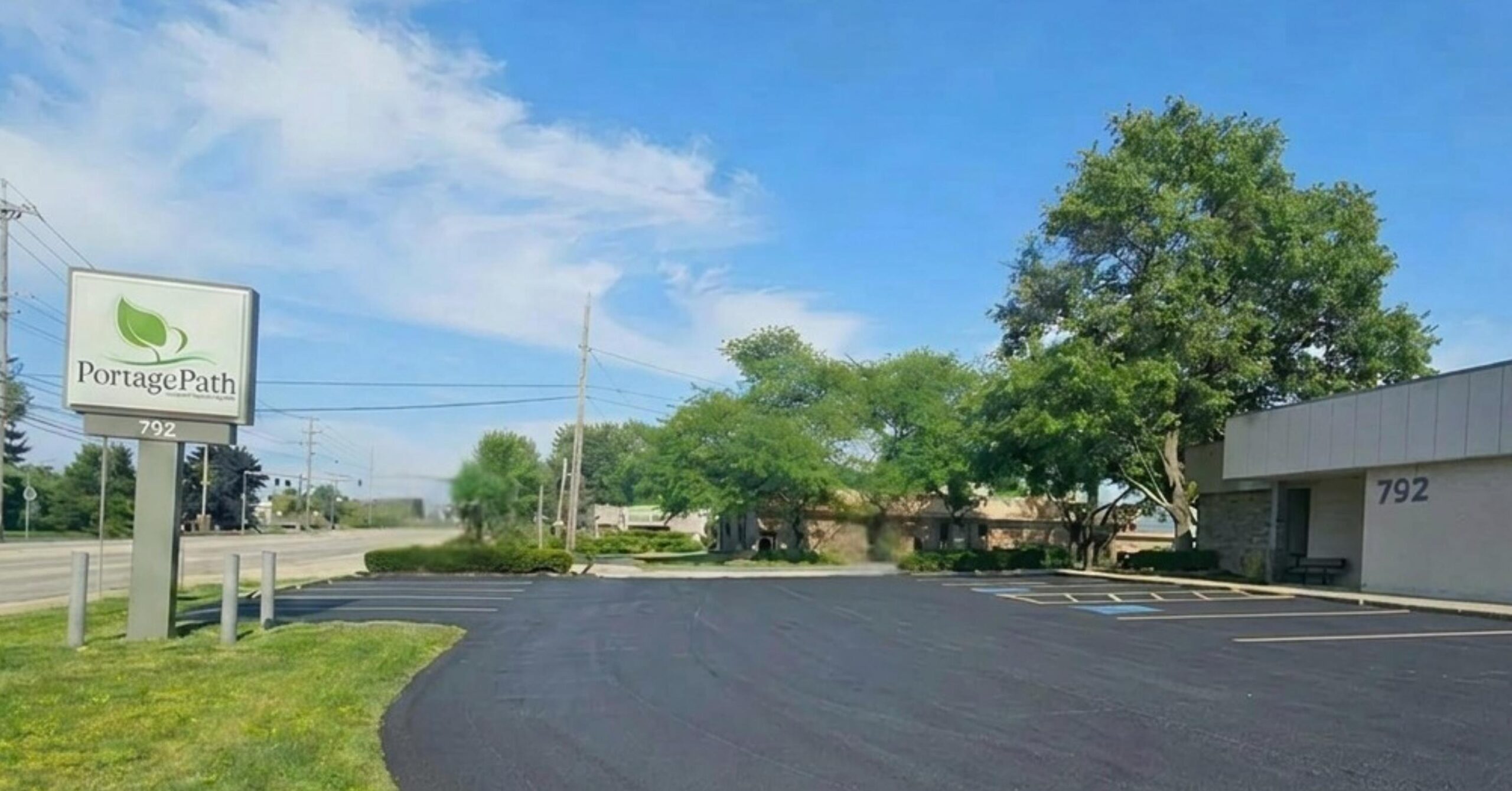 North Summit Parking Lot Photo | Portage Path Behavioral Health Portage path sign with green leaf logo beside a shaded parking lot and a building marked 792 against a blue sky.