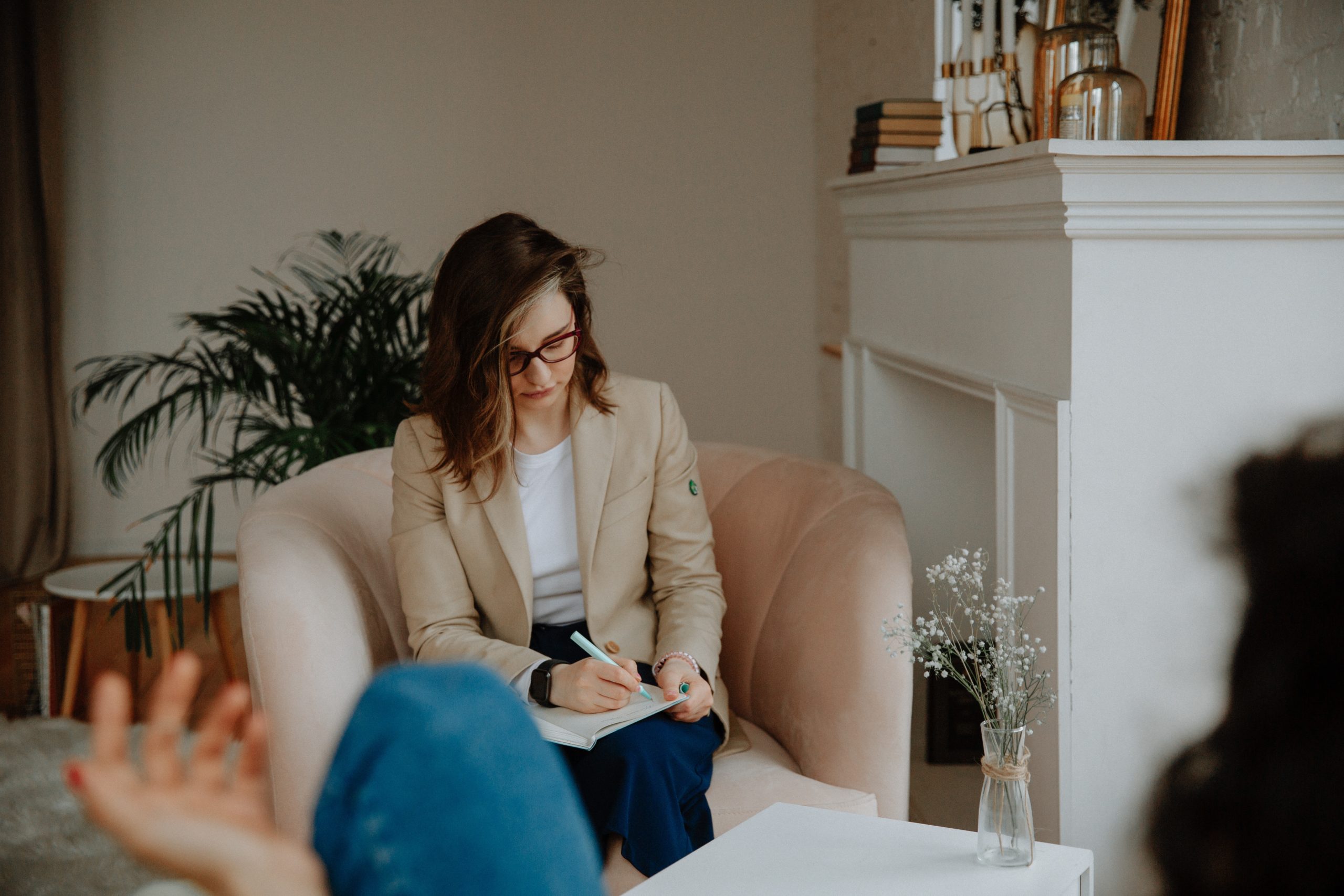 Outpatient-hero-image | Portage Path Behavioral Health Woman with glasses sits on a beige sofa, writing in a notebook, as blurred hands appear in the foreground.