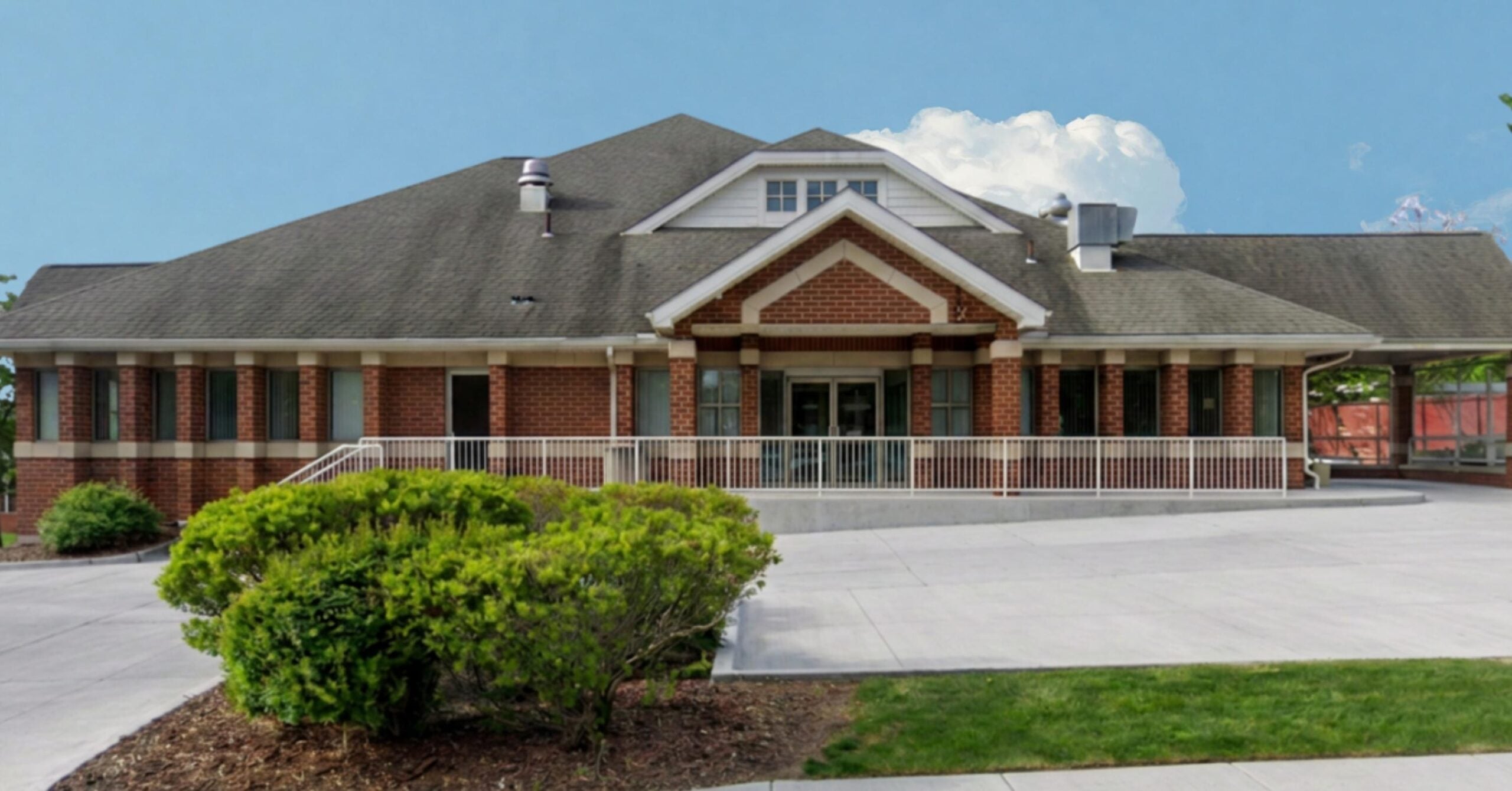 Pes Exterior | Portage Path Behavioral Health Brick, single-story commercial building with a central covered entrance, white railing, and brick columns; landscaping in front and a blue sky above.
