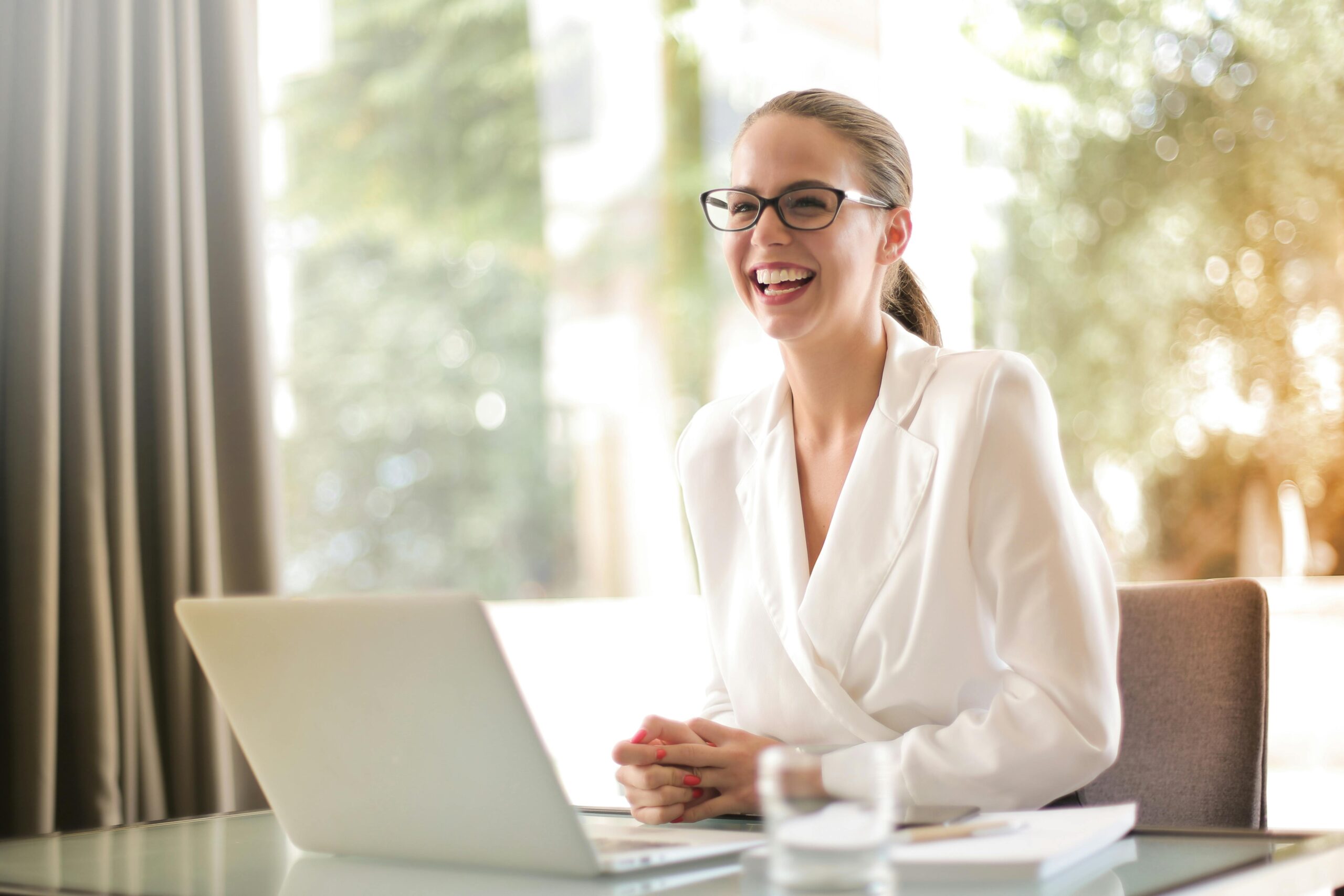 Smilingbusinesswoman - Portage Path Behavioral Health Smiling businesswoman with glasses working on a laptop at a glass table in a bright office setting.