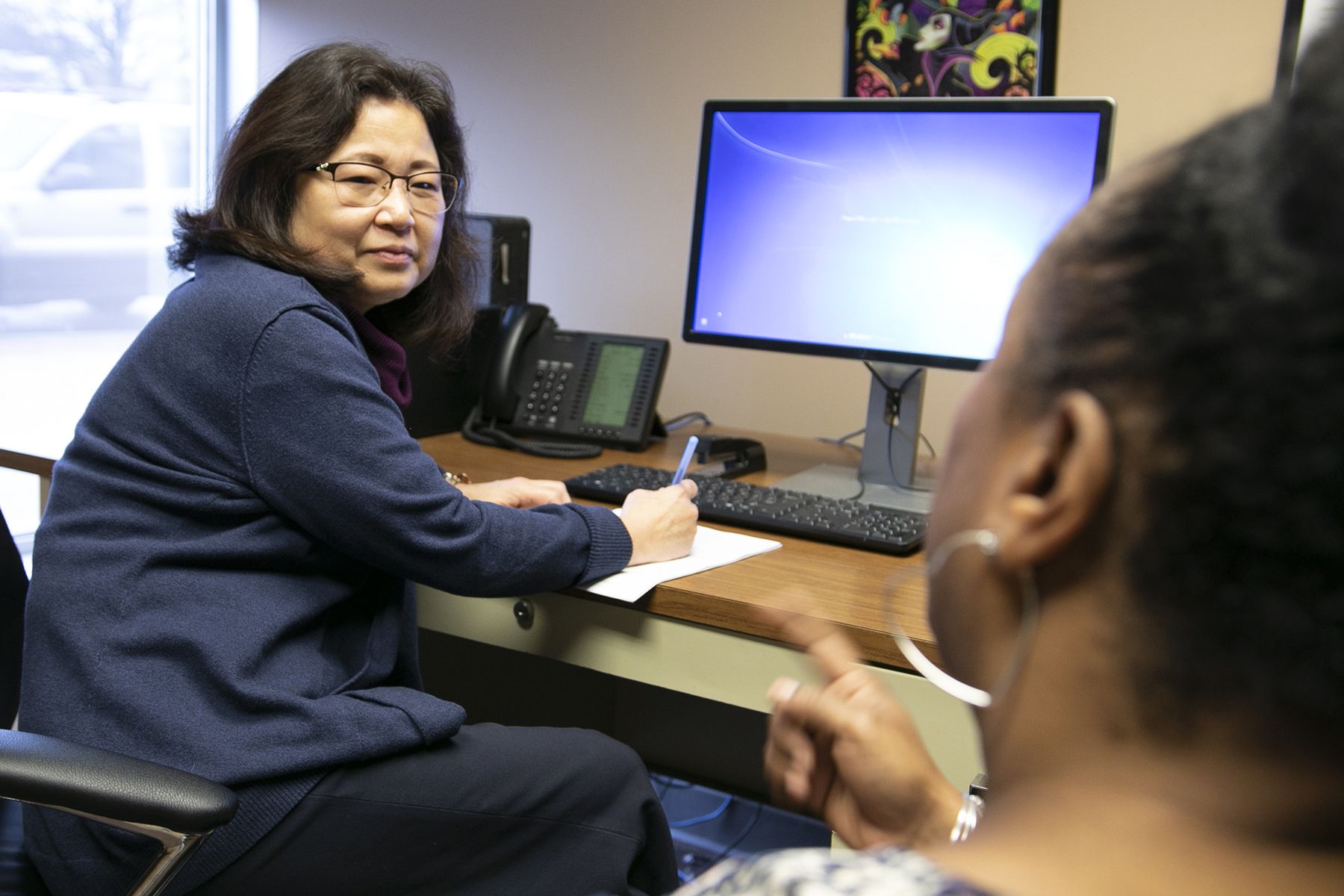 Woman in a blue sweater sits at a desk, writing on paper as a computer monitor and office phone are nearby in a professional office setting.