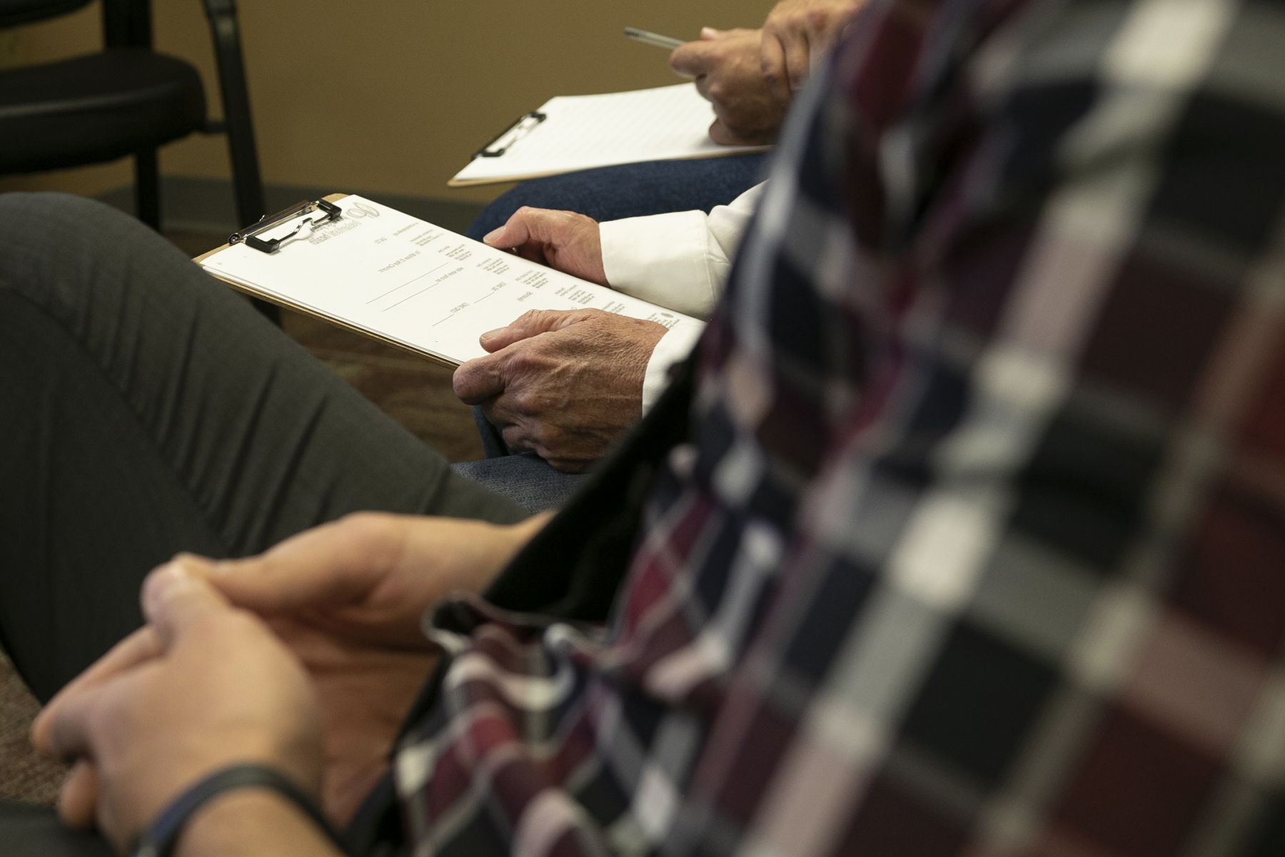 Two people are seated; one is writing on a clipboard while the other holds a document, suggesting a form or discussion in a waiting/office setting.